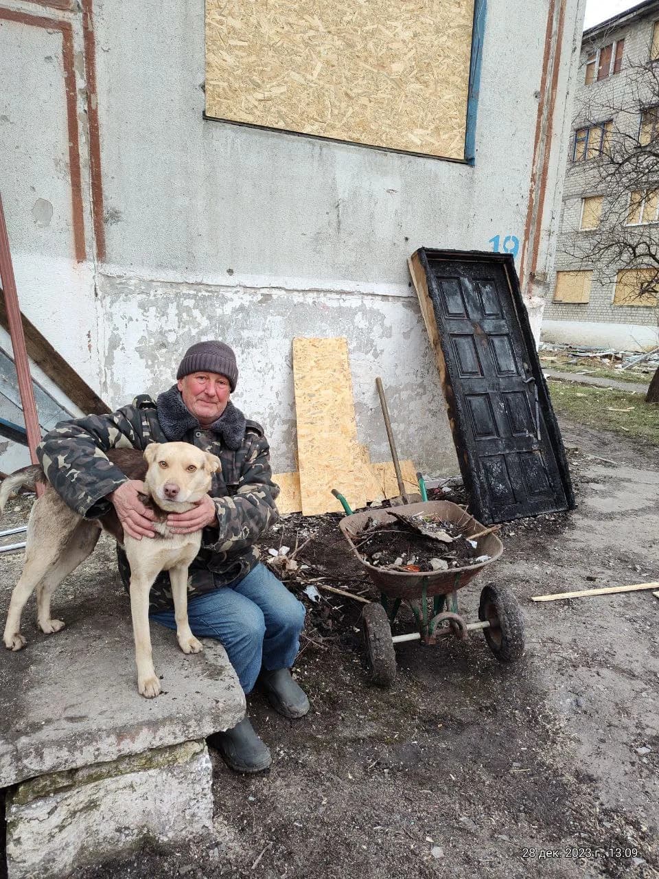 Відновлення пошкодженого житла. Курилівка, військове містечко, грудень 2024 року. На фото один із жителів, колишній військовий, полковник Повітряних сил України у відставці. Він в обіймах із собакою біля своїх дверей, квартири та закритого вікна.