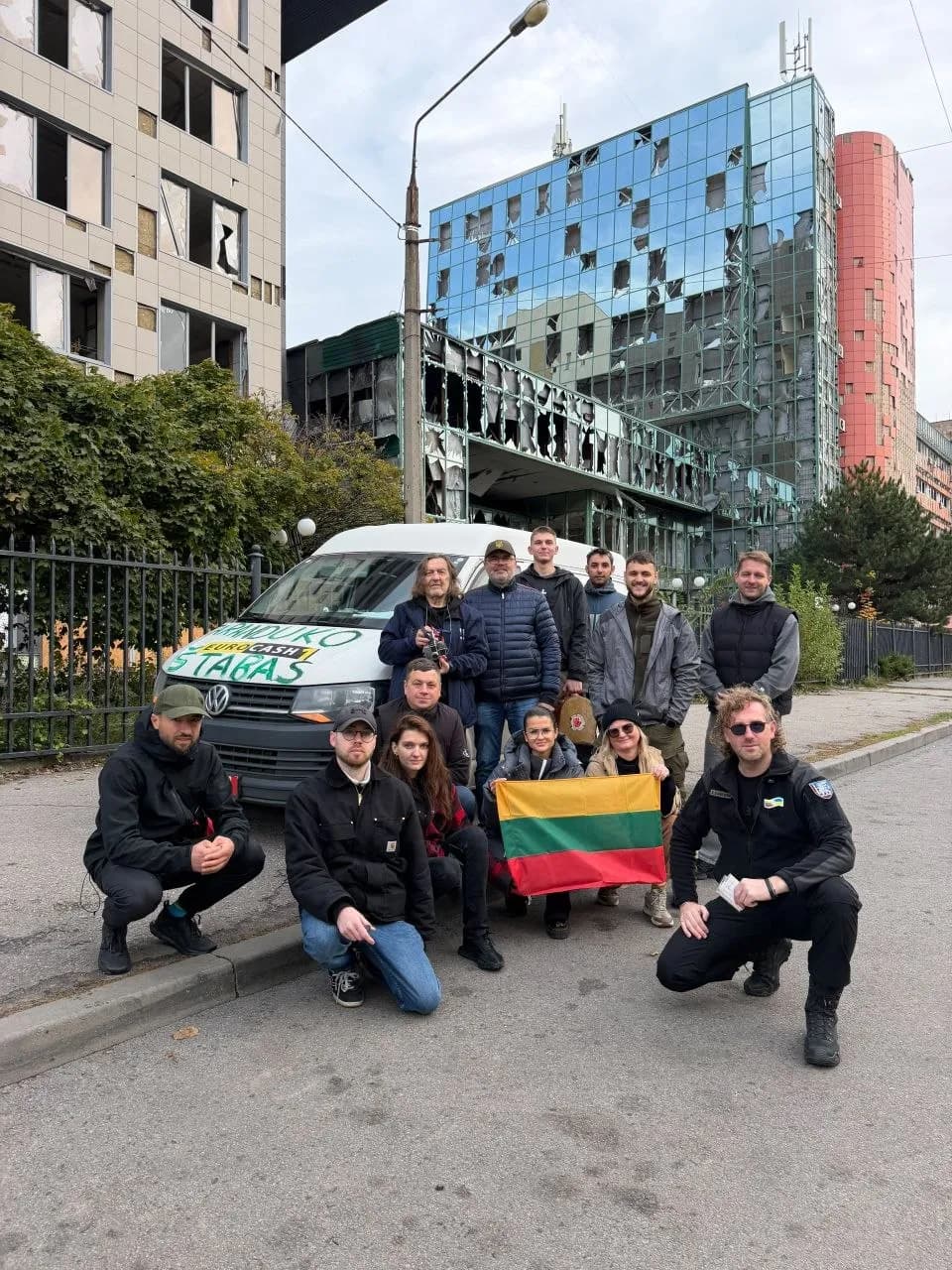 группа людей с литовским флагом возле белого микроавтобуса на фоне повреждённых зданий с выбитыми окнами